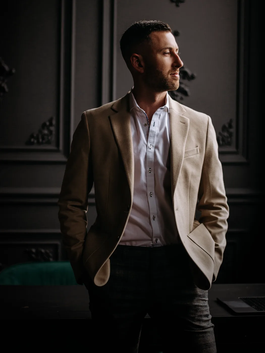 Kurt Casucci in a cream blazer and white shirt, standing against a dark panelled wall with a laptop on the desk behind him